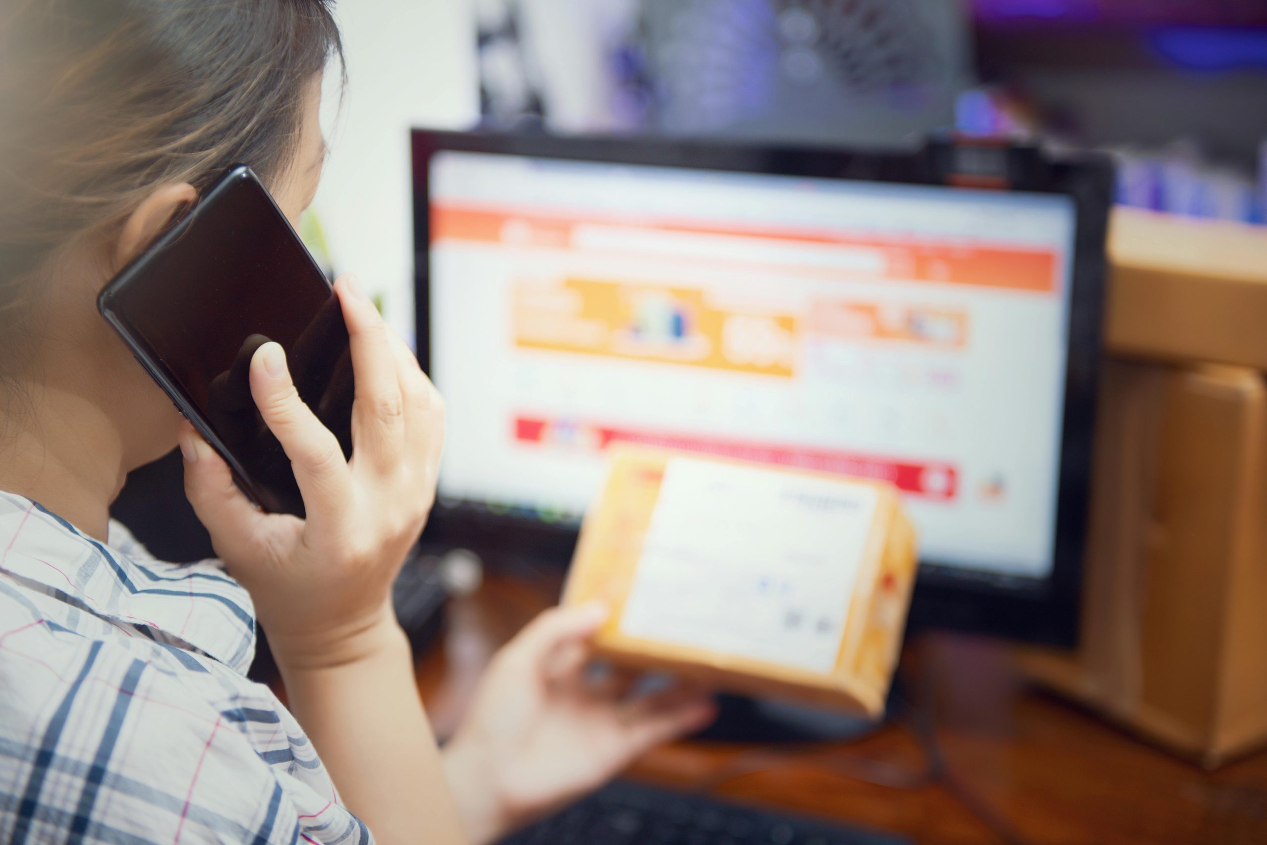Customer on the phone holding a parcel while viewing an online order page on a computer, suggesting a delivery or tracking enquiry
